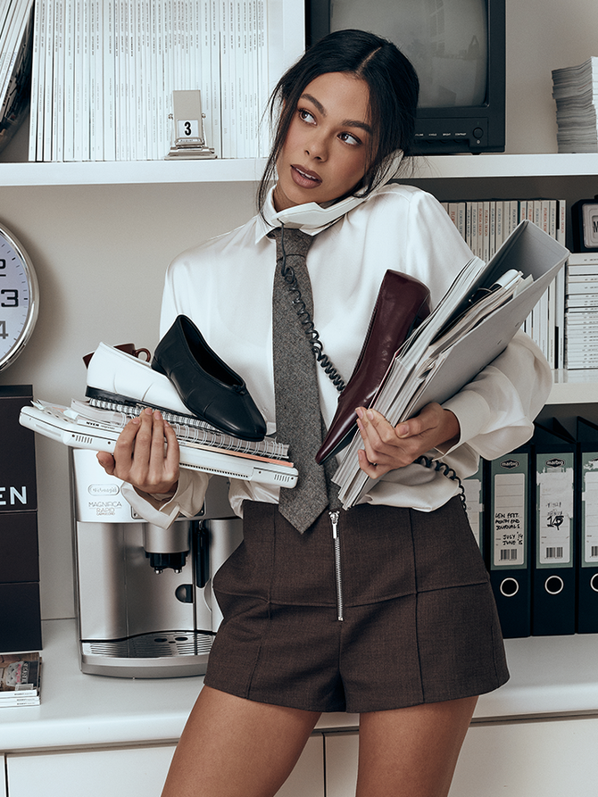 A woman in an office setting wearing a white blouse, gray tie, and brown shorts, holding a stack of books, shoes, and folders.