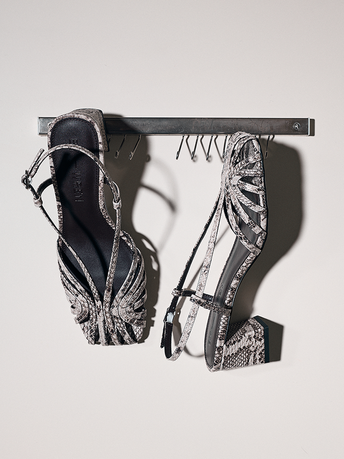 A pair of women's strappy heeled sandals with a snakeskin pattern, hanging from a black metal rack against a light gray wall.