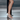 White strappy high-heeled sandals worn on a model's feet, shown in a close-up shot against a metallic gray background.